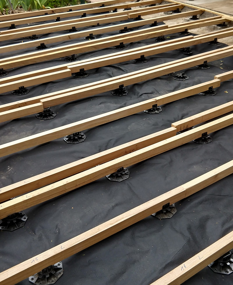 Terrassen-Unterkonstukrionshölzer aus Eiche auf Stellfüßen platziert, darunter eine Unkrautschutzfolie.